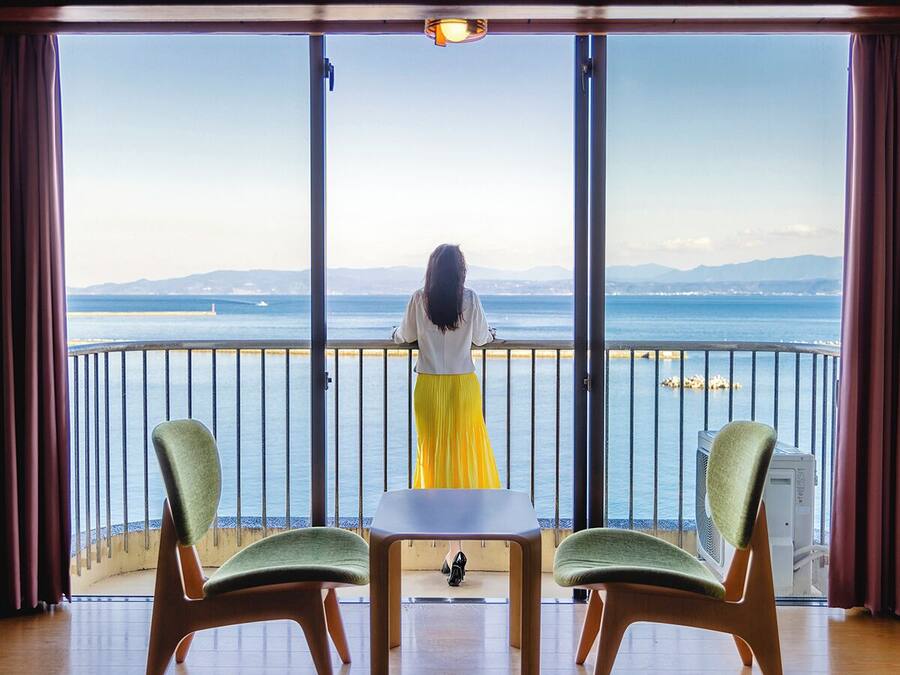 Un balcon donnant sur la mer, une table, deux chaises et une personne se tenant près de la balustrade.