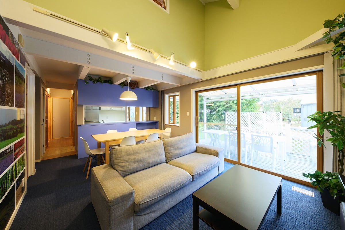 A modern living room with a sofa, a coffee table, and a dining area with a table and chairs. There is a kitchen area with a blue countertop and a window overlooking a garden.