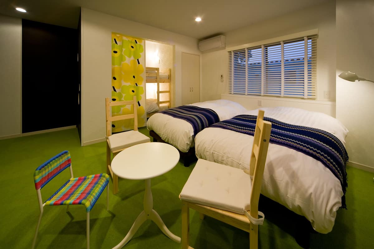 A room with two beds, a small table, a chair, a bunk bed, and a window with blinds.