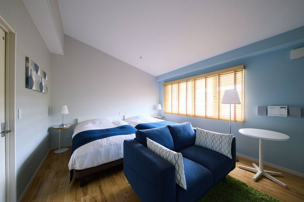 A bedroom with a bed, a blue sofa, a small round table, and a window with blinds.
