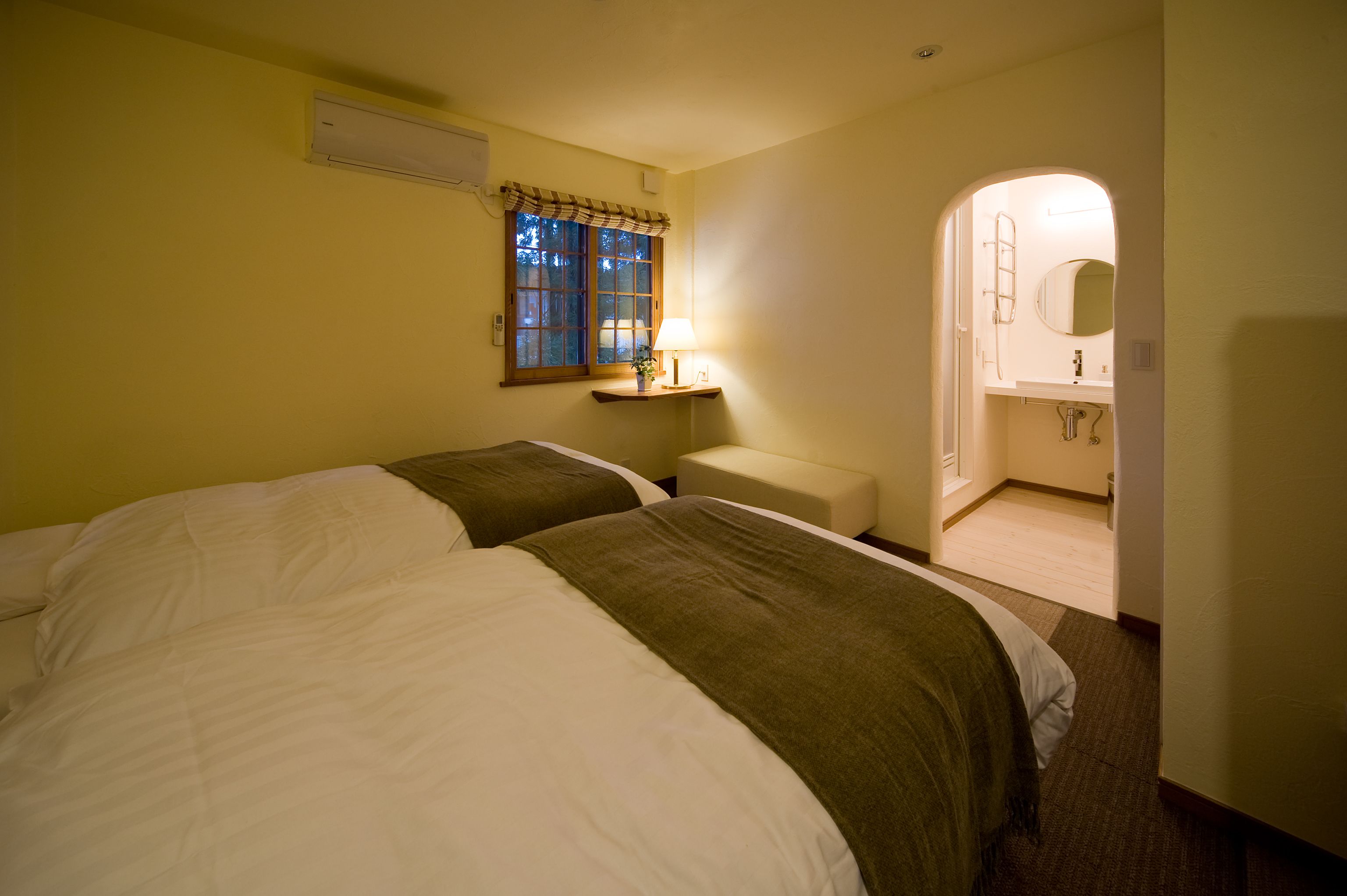 Quarto de hotel com duas camas, uma mesa de cabeceira com abajur, uma janela com cortinas e um banheiro visível através de um arco.