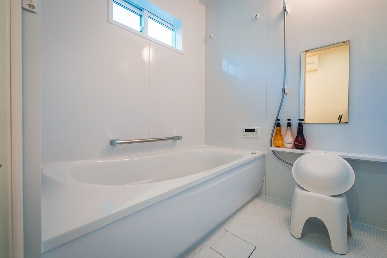 A modern bathroom with a white bathtub, a small stool, a shelf with toiletries, and a window above the tub.