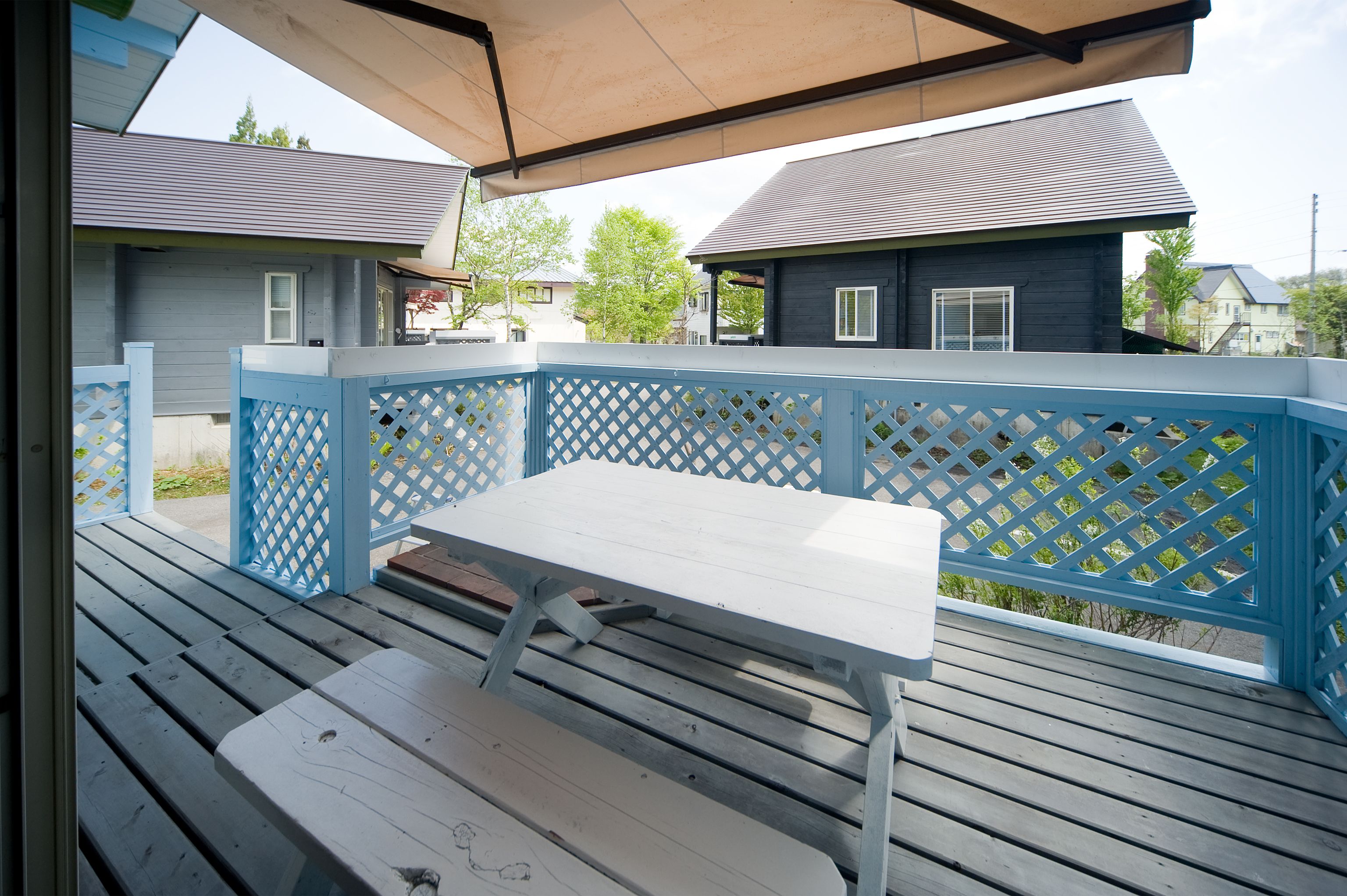 A covered outdoor deck with a wooden table and benches, surrounded by a blue lattice fence.