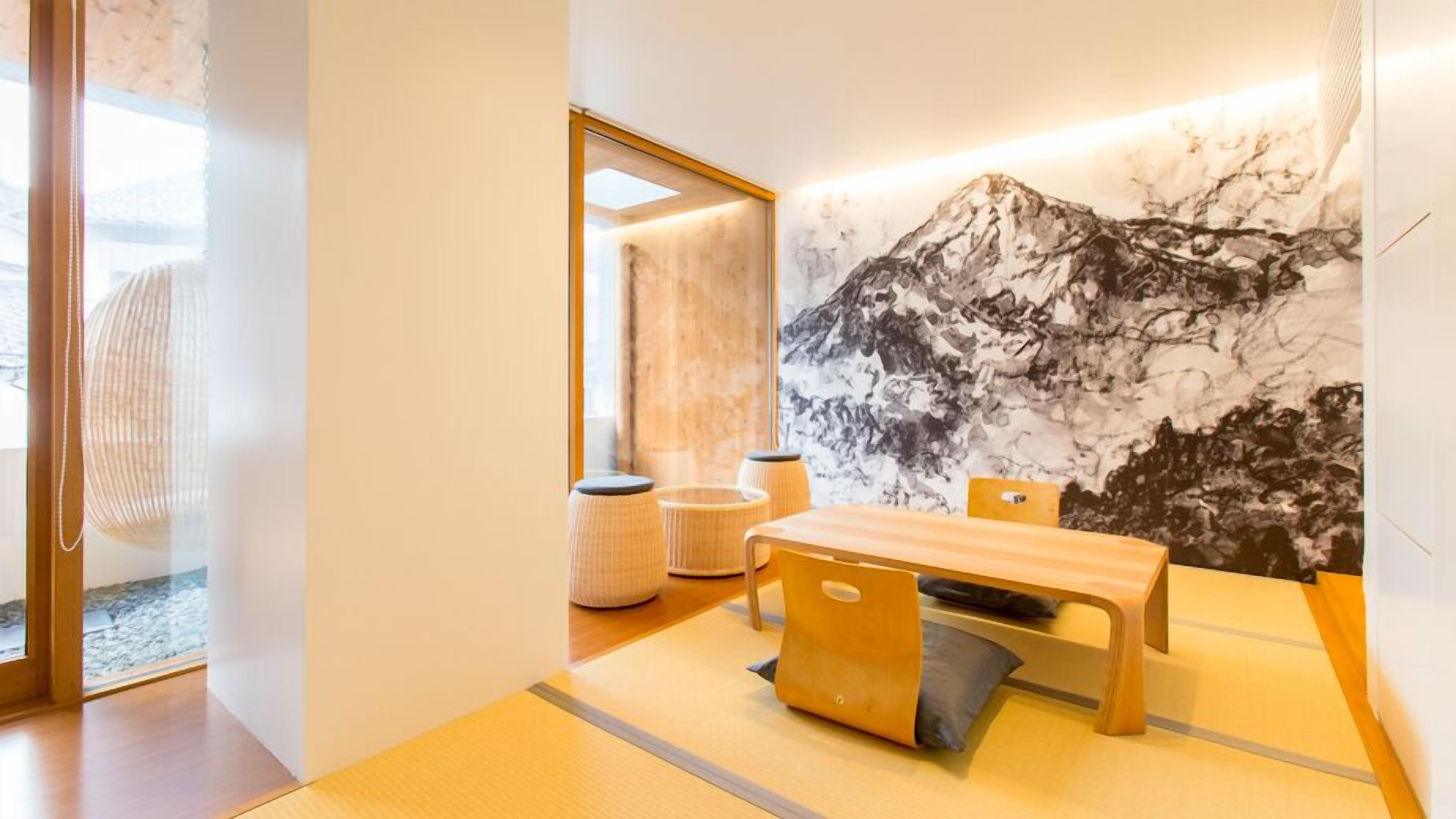 Una habitación con una mesa de madera, sillas, un cuadro grande de una montaña y un ventanal de vidrio que da al exterior.