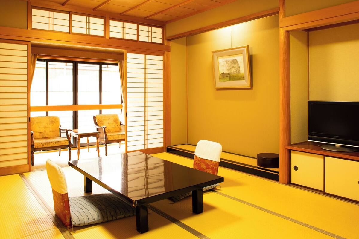 Een traditionele Japanse kamer met tatami vloerbedekking, een lage tafel, twee stoelen, een televisie en schuifdeuren.