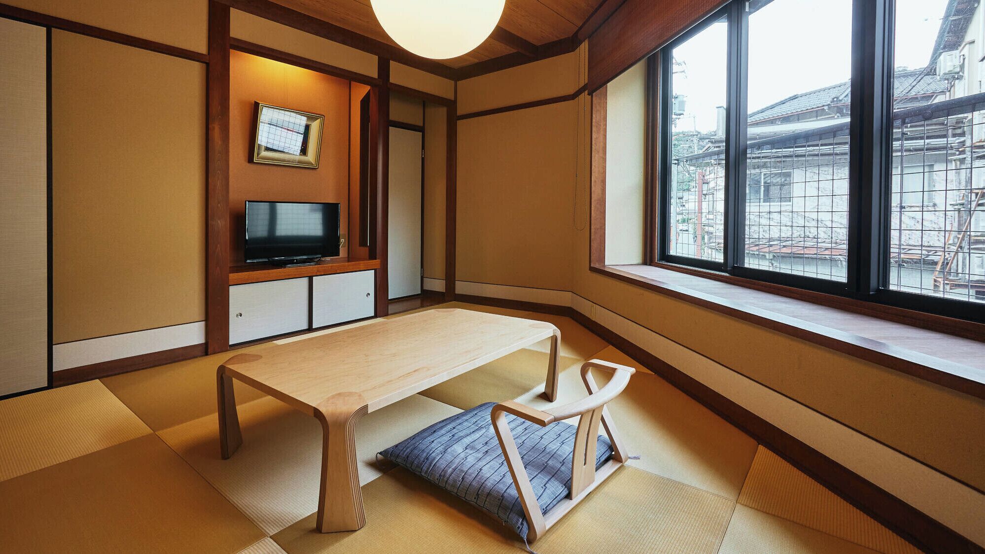 Una habitación japonesa tradicional con suelo de tatami, una mesa de madera baja, un televisor y una ventana con vistas a edificios.