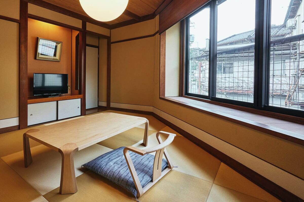 Een traditionele Japanse kamer met tatami vloeren, een lage houten tafel, een televisietoestel en een raam met uitzicht op gebouwen.