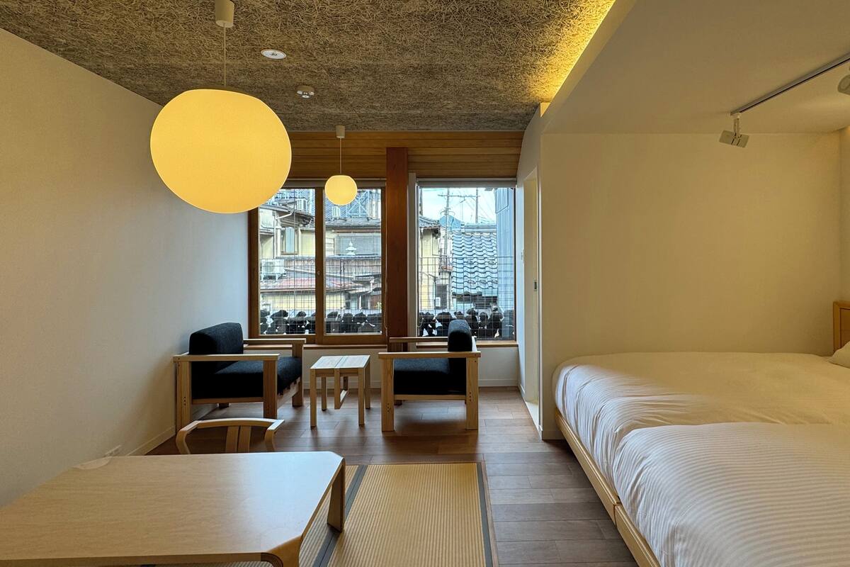 Een hotelkamer met een bed, een bureau, twee stoelen, een raam met uitzicht op gebouwen, en een plafond met een structuur.
