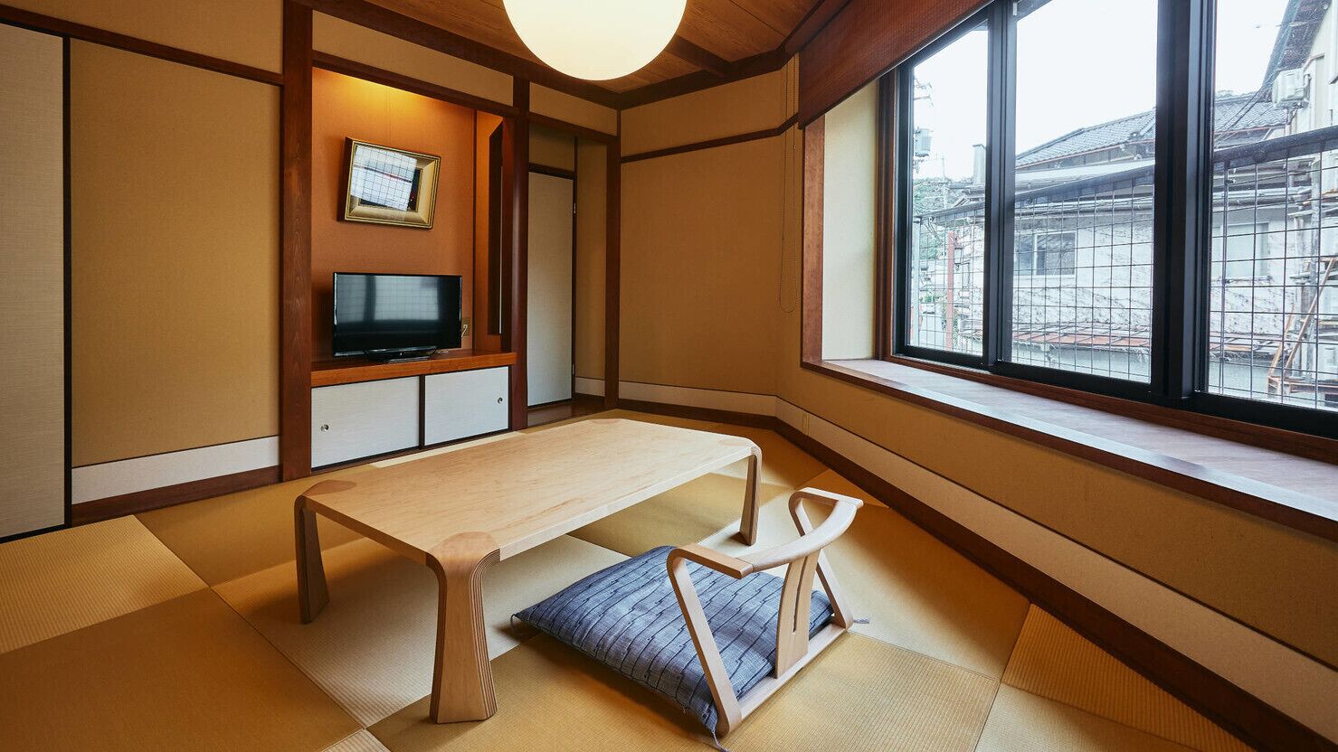 Una habitación japonesa tradicional con suelo de tatami, una mesa de madera baja, un televisor y un ventanal con vistas a edificios.