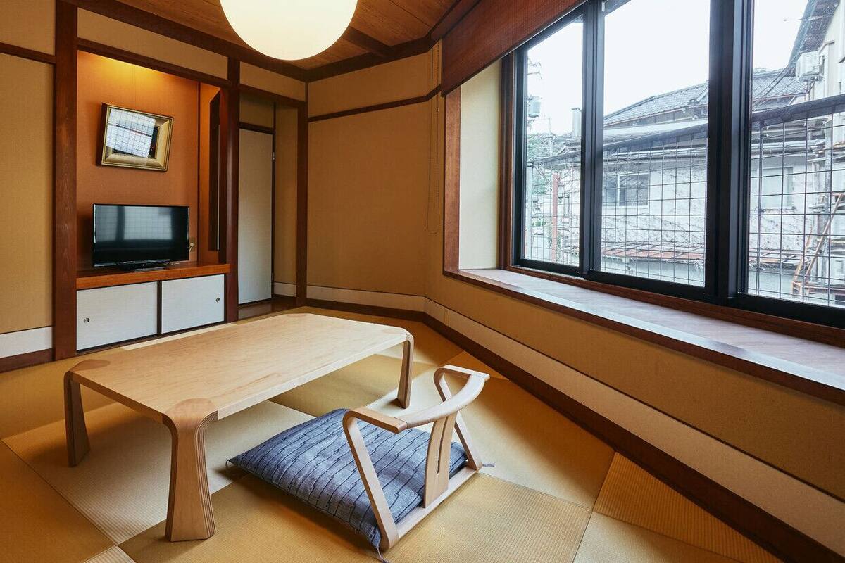 Een traditionele Japanse kamer met tatami vloerbedekking, een lage houten tafel, een televisie en een raam met uitzicht op gebouwen.