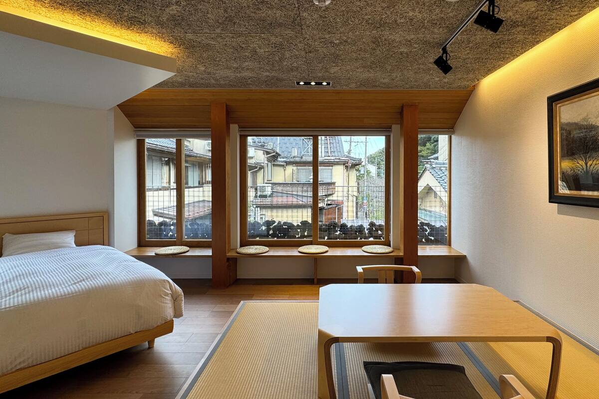 Een hotelkamer met een bed, een tafel, een stoel, een raam met uitzicht op gebouwen en een schilderij aan de muur.