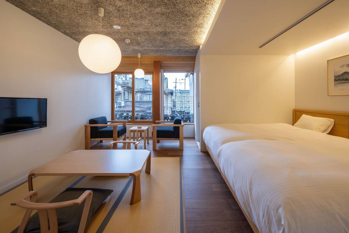 Een moderne hotelkamer met een bed, een televisie, een eettafel, stoelen en uitzicht op gebouwen door grote ramen.