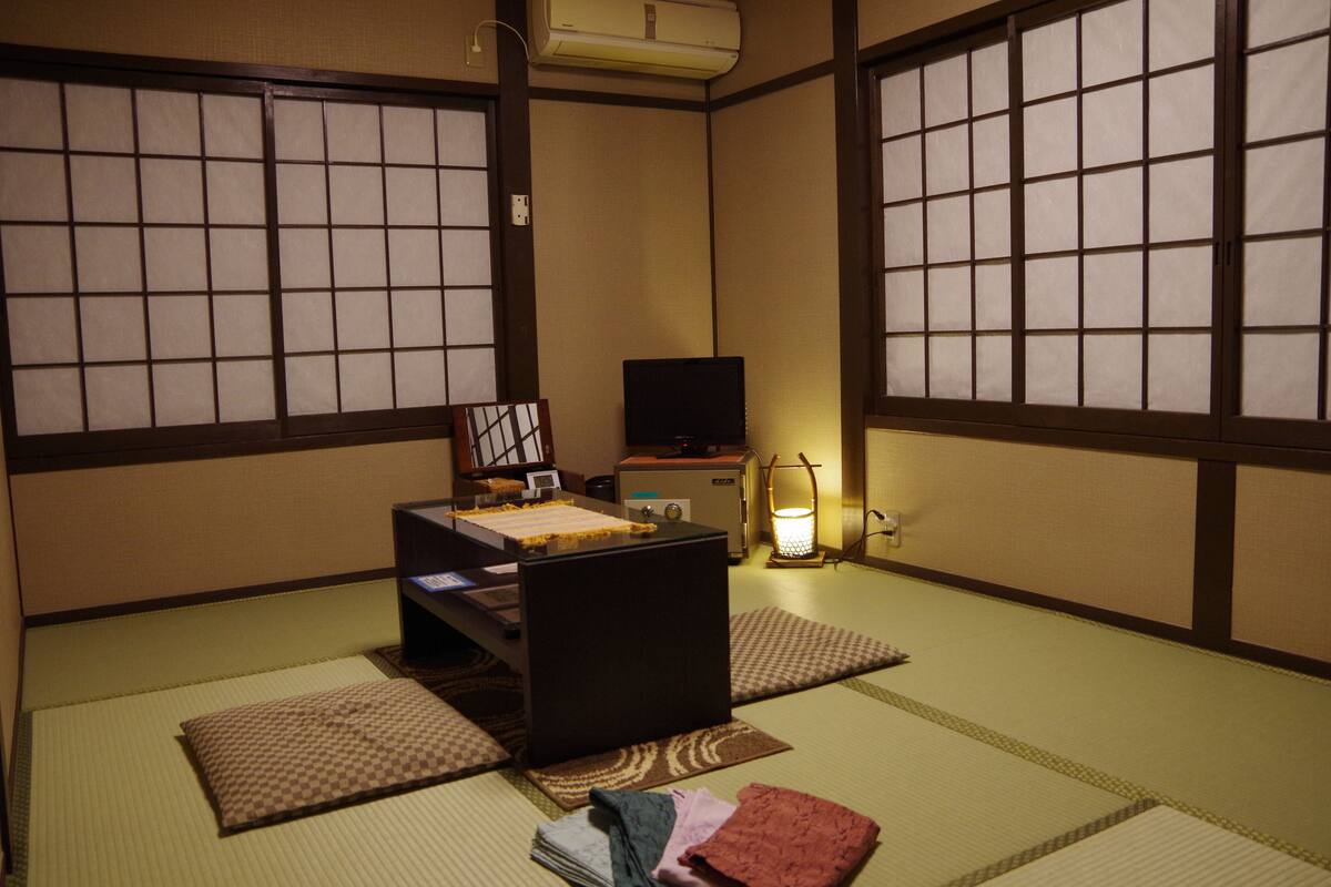 Ein traditioneller japanischer Raum mit Tatami-Boden, einem niedrigen Tisch mit Kissen, einem Fernseher und einer Nachttischlampe.