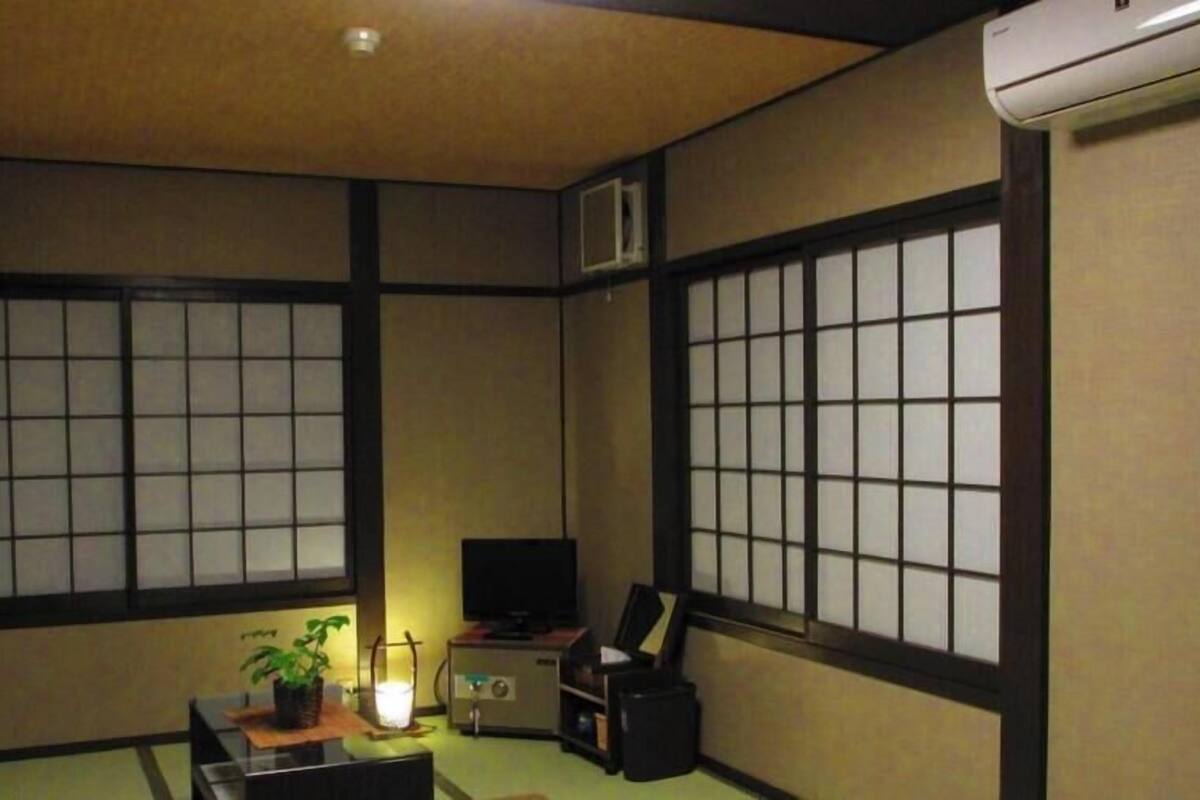 Ein traditioneller japanischer Raum mit Tatami-Boden, Shoji-Schirmwänden, einem Fernseher, einem Schreibtisch mit Computer und einer kleinen Pflanze.