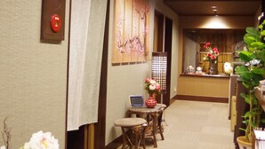 Un couloir d’hôtel avec un comptoir de réception, un coin salon équipé d’un ordinateur portable, et des éléments décoratifs tels que des fleurs et des œuvres d’art mural.