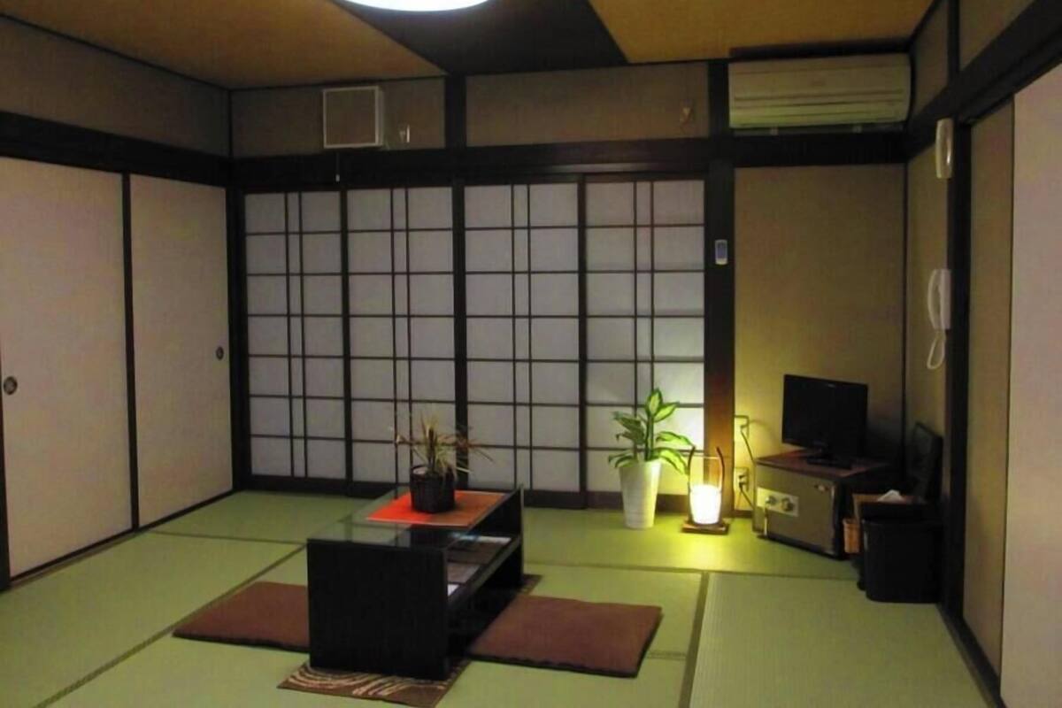 Ein traditioneller japanischer Raum mit Tatami-Boden, einem niedrigen Tisch mit Pflanze, einem Fernseher und einem Heizgerät.