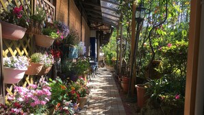 Un étroit sentier bordé de plantes et de fleurs en pot, qui mène à un espace abrité avec une table et des chaises.