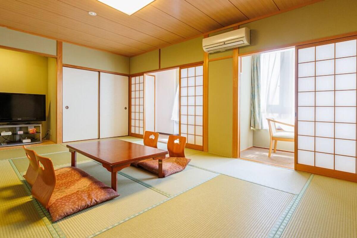 A traditional Japanese room with tatami flooring, a low table with cushions, sliding doors, and a television.