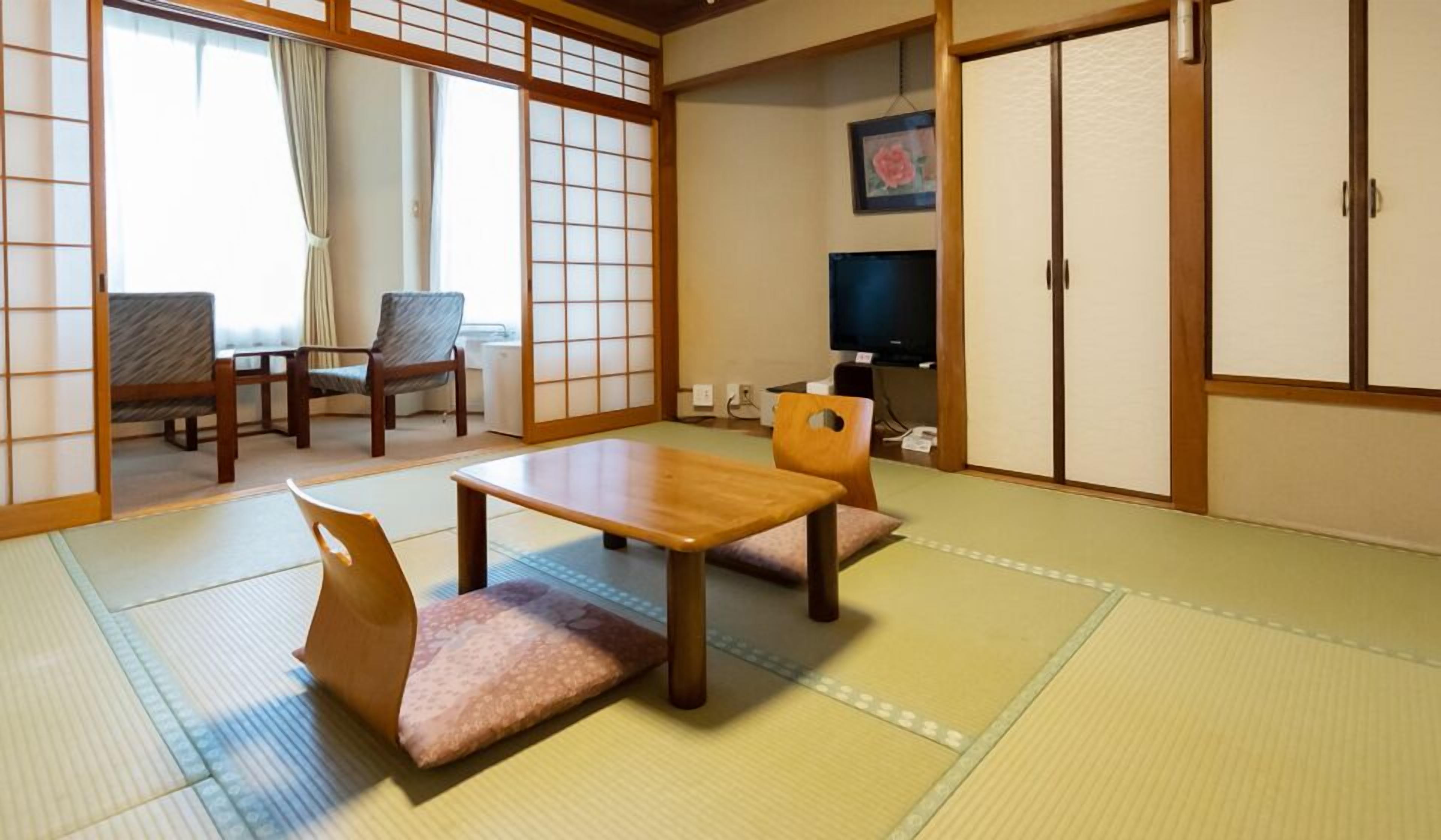 Une pièce de style japonais traditionnel, avec un sol recouvert de tatamis, une table basse, deux chaises, un téléviseur et des portes coulissantes.