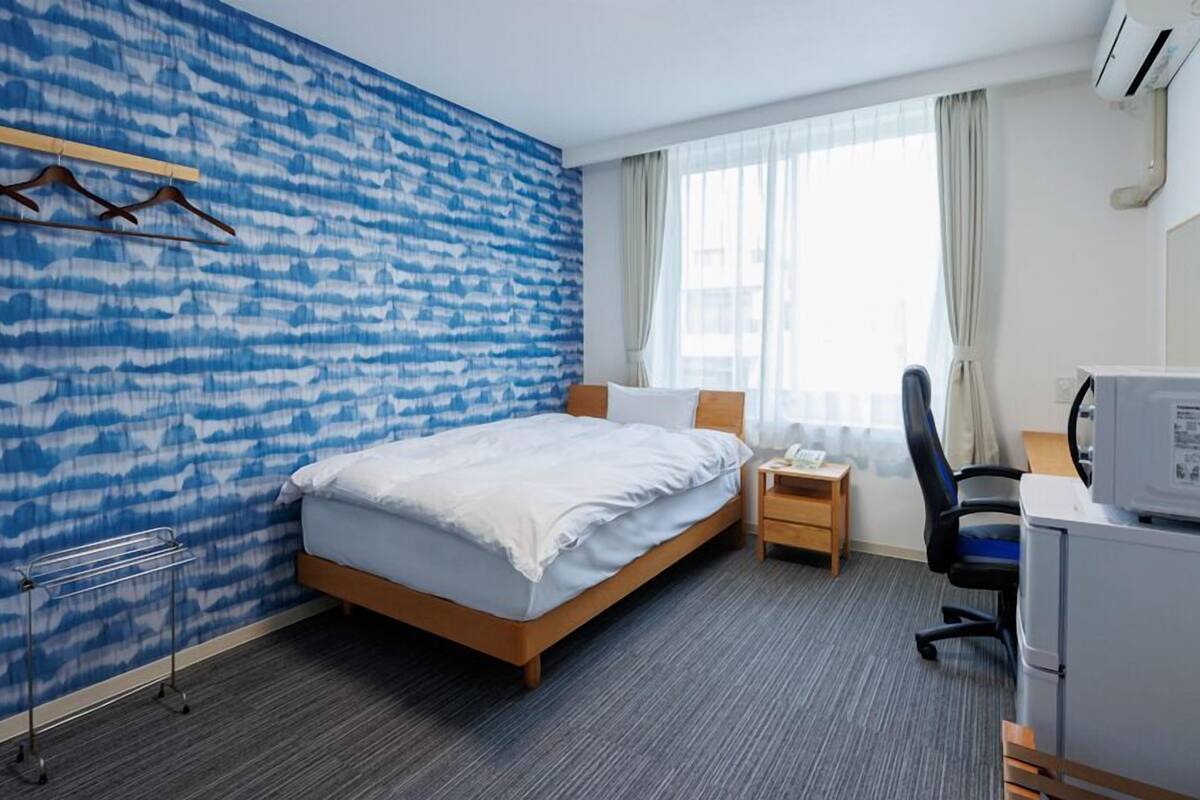 A hotel room with a bed, a desk, a chair, a small table, and a wall-mounted air conditioner.