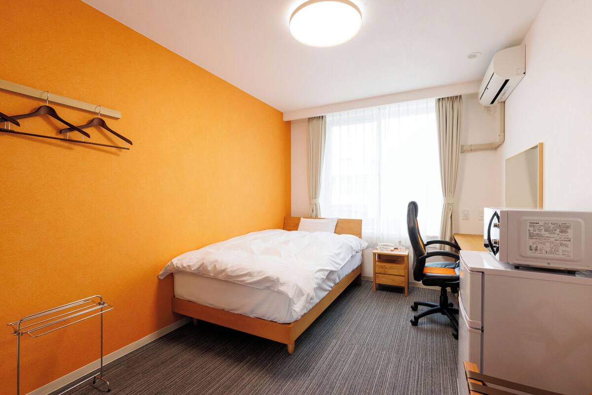 A hotel room with a bed, a desk with a computer, a chair, a radiator, and a window with curtains.
