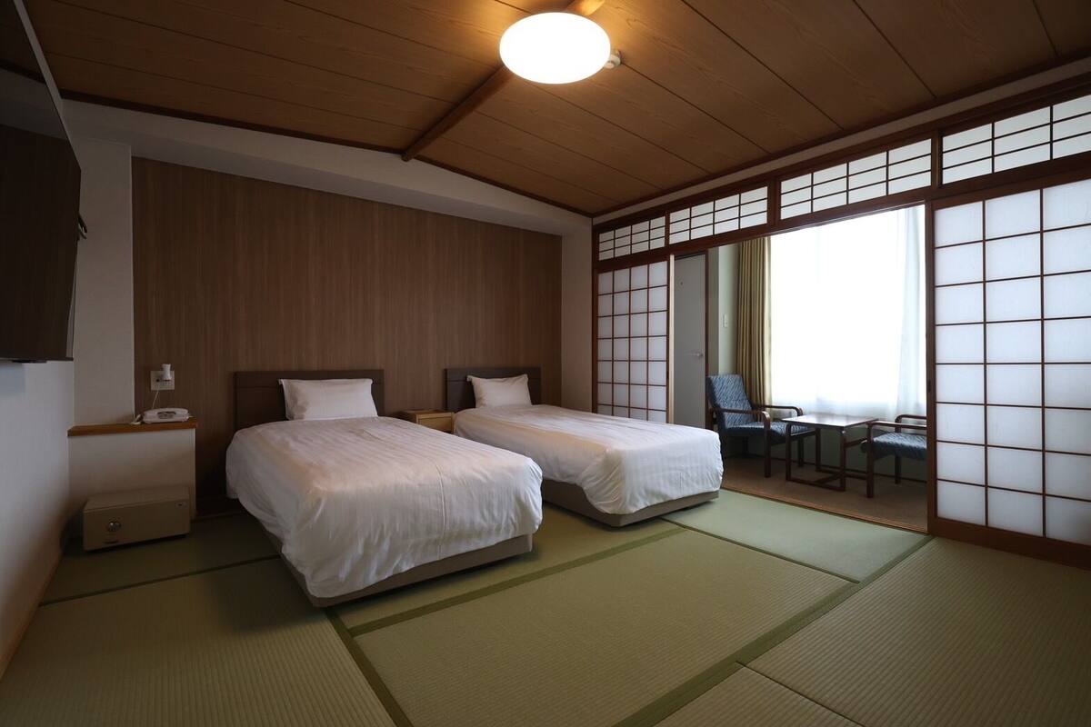 A traditional Japanese room with tatami flooring, two beds, a desk with chairs, and a sliding shoji screen.