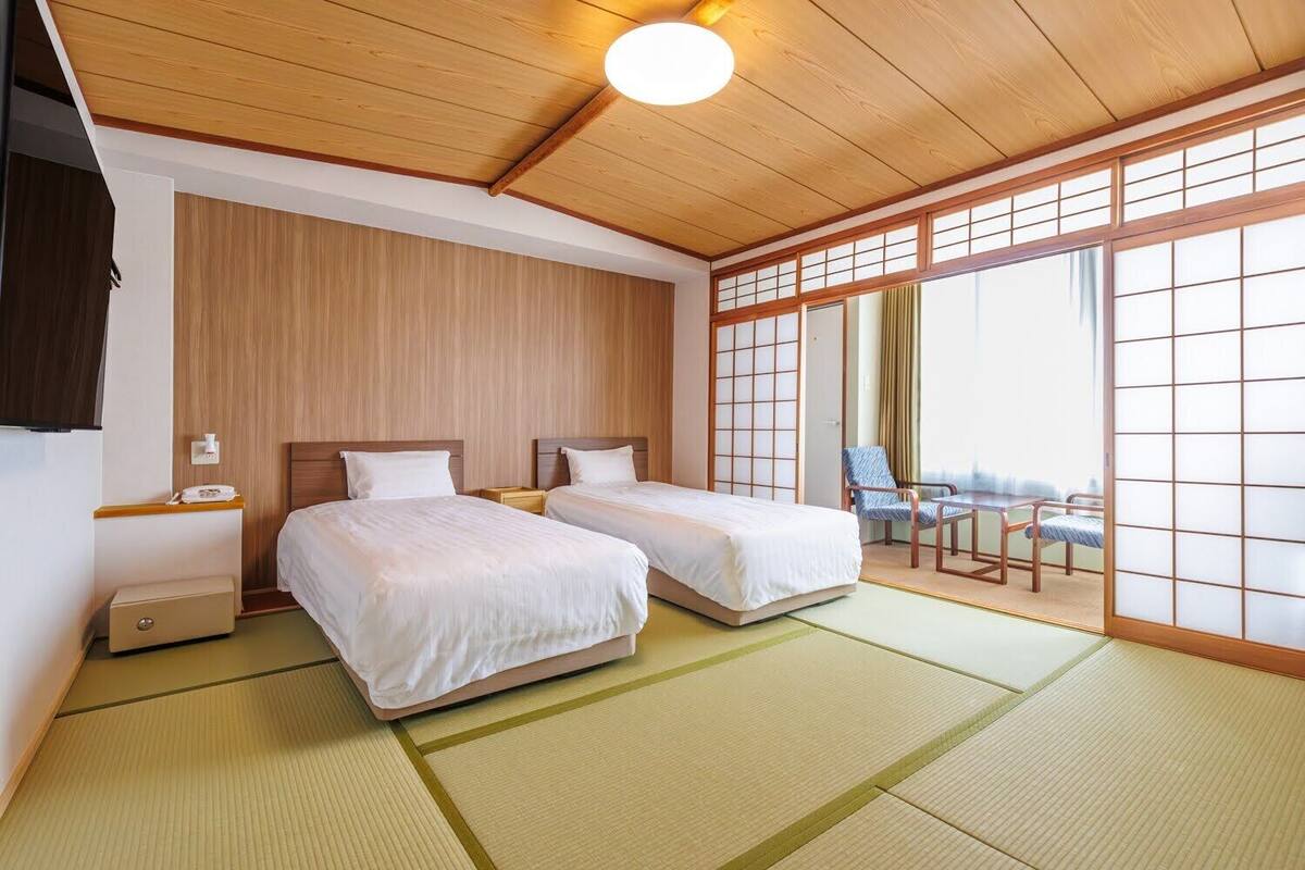 A hotel room with two beds, a wooden ceiling, a sliding shoji screen, tatami mats, a small table, and a chair.