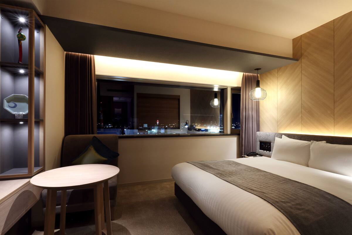 A hotel room with a bed, a desk, a chair, a window with a city view, and a nightstand with a lamp.