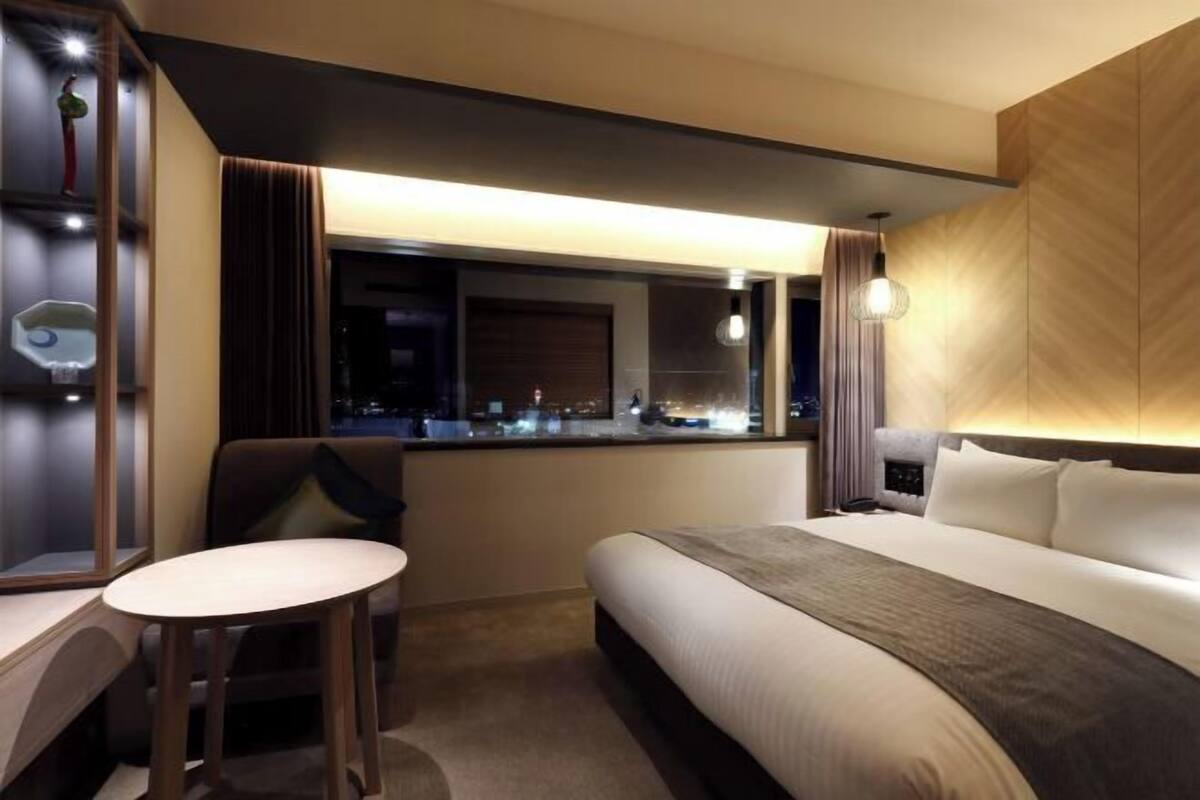 A hotel room with a bed, a small table, a chair, a nightstand, a mirror, and a window with a view of the city at night.