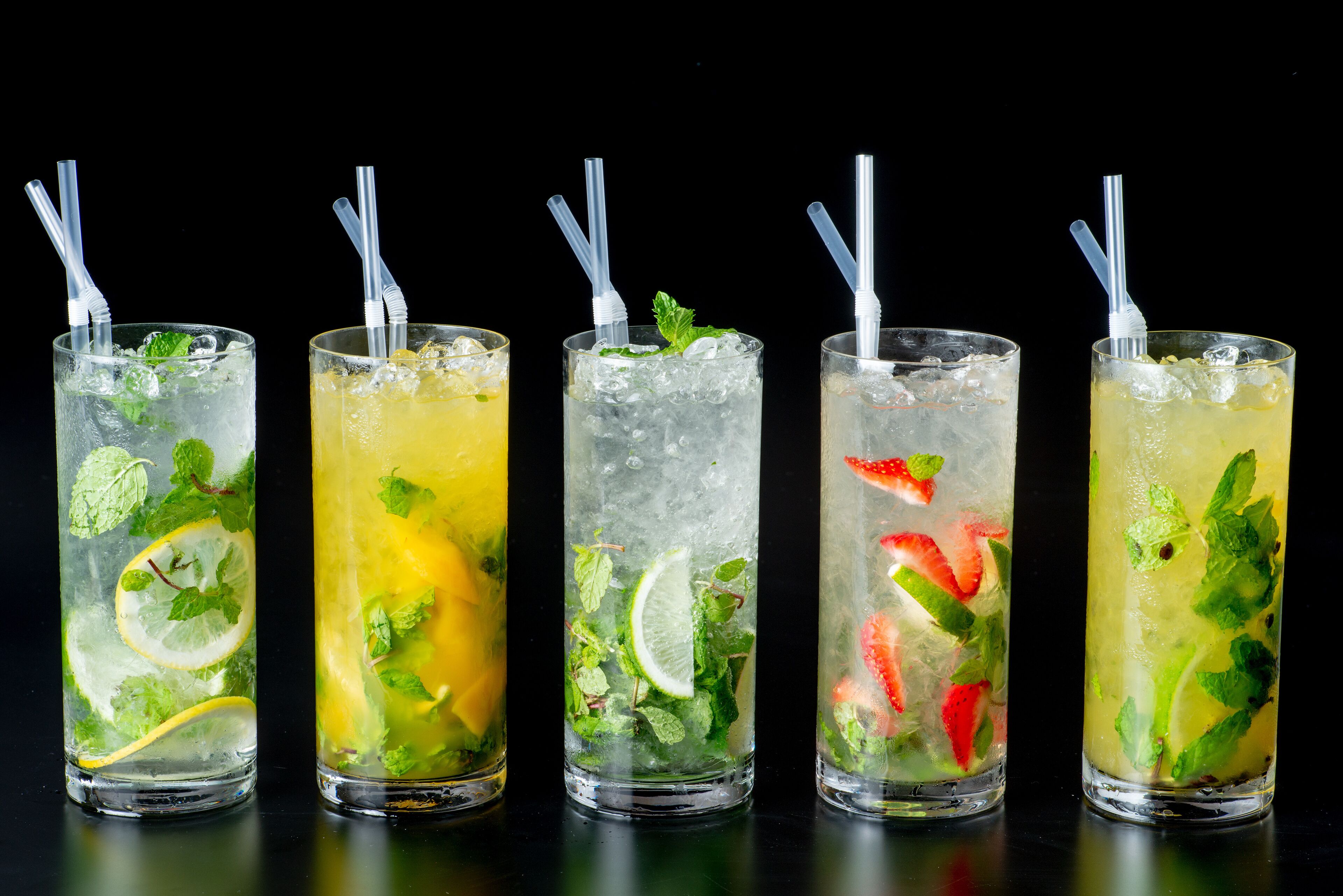 Cinco vasos altos de bebidas heladas con pajitas, adornados con hojas de menta y rodajas de cítricos.