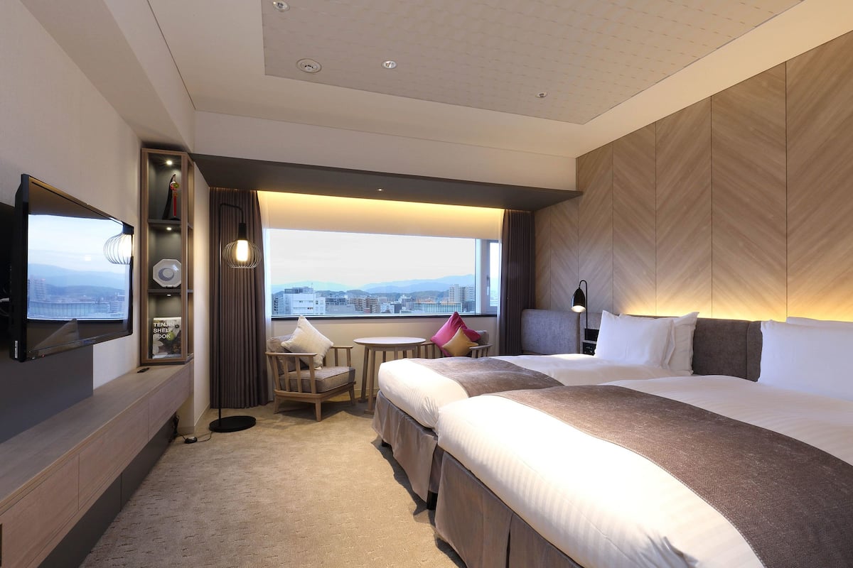 A hotel room with two beds, a TV, a small table, and a view of the city.