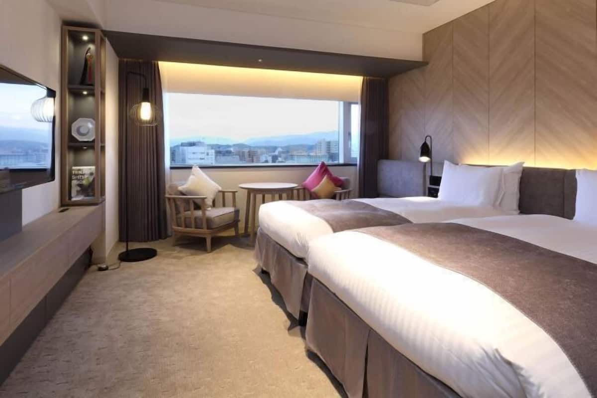A hotel room with two beds, a desk, a chair, a small table, a TV, and a view of the city.