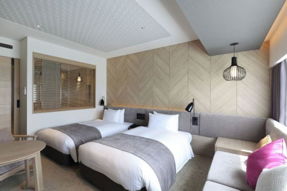 Una habitación de hotel moderna con dos camas, un sofá, una mesa de comedor y un ventanal con persianas.