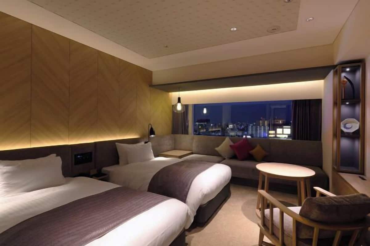 A hotel room with two beds, a sofa, a small table, and a city view.