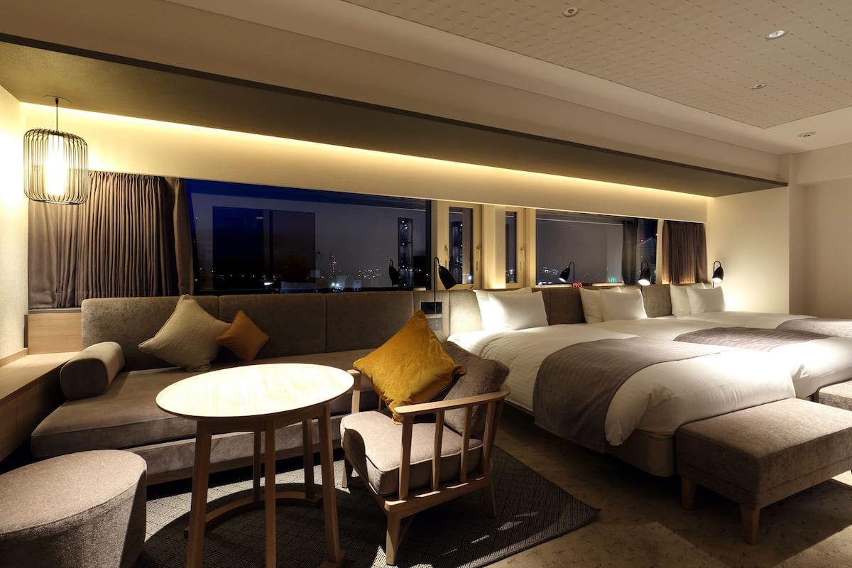 A modern hotel room with a sofa, a round table, a bed with a large bedspread, and a view of the city at night.