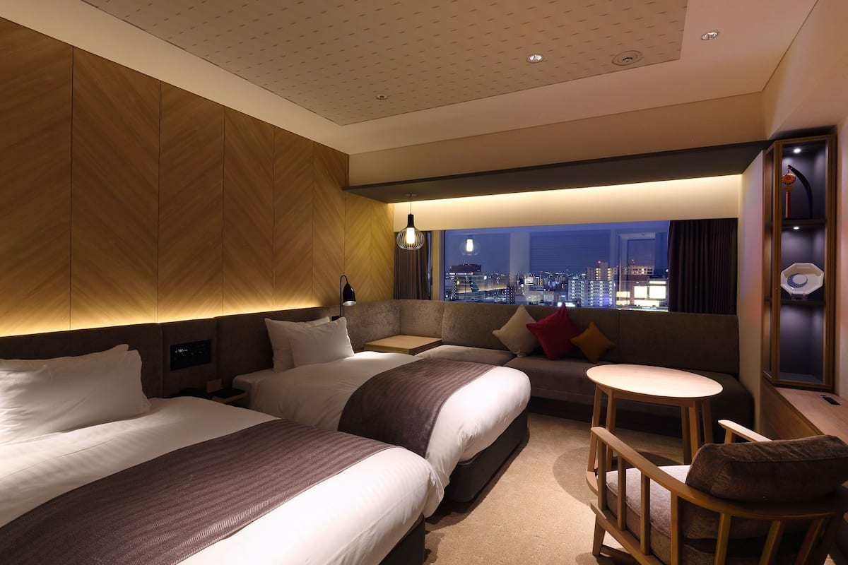 A modern hotel room with two beds, a sofa, a small table, and a city view.
