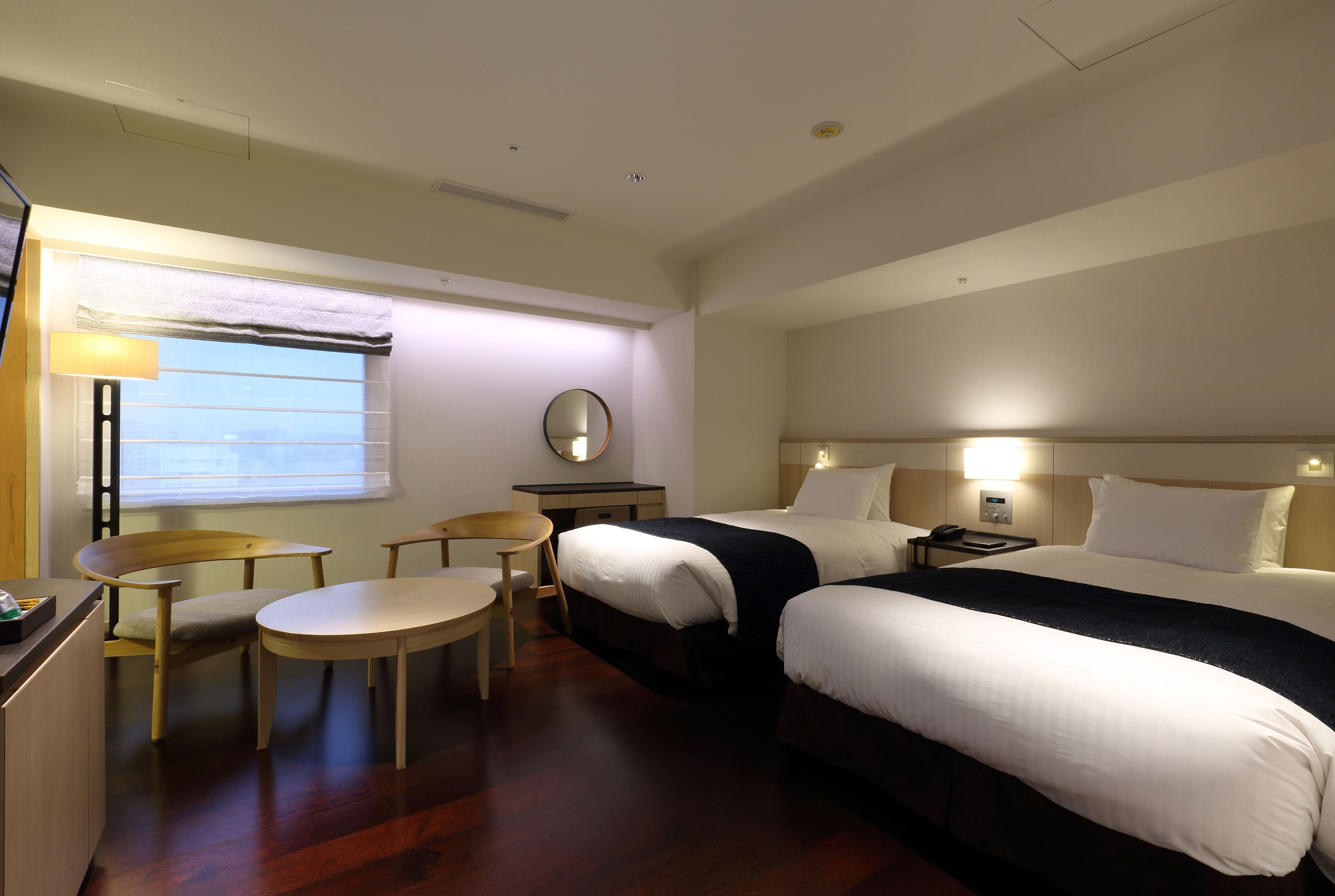 Quarto de hotel com duas camas, uma escrivaninha, uma cadeira, uma mesinha e uma janela com persianas.