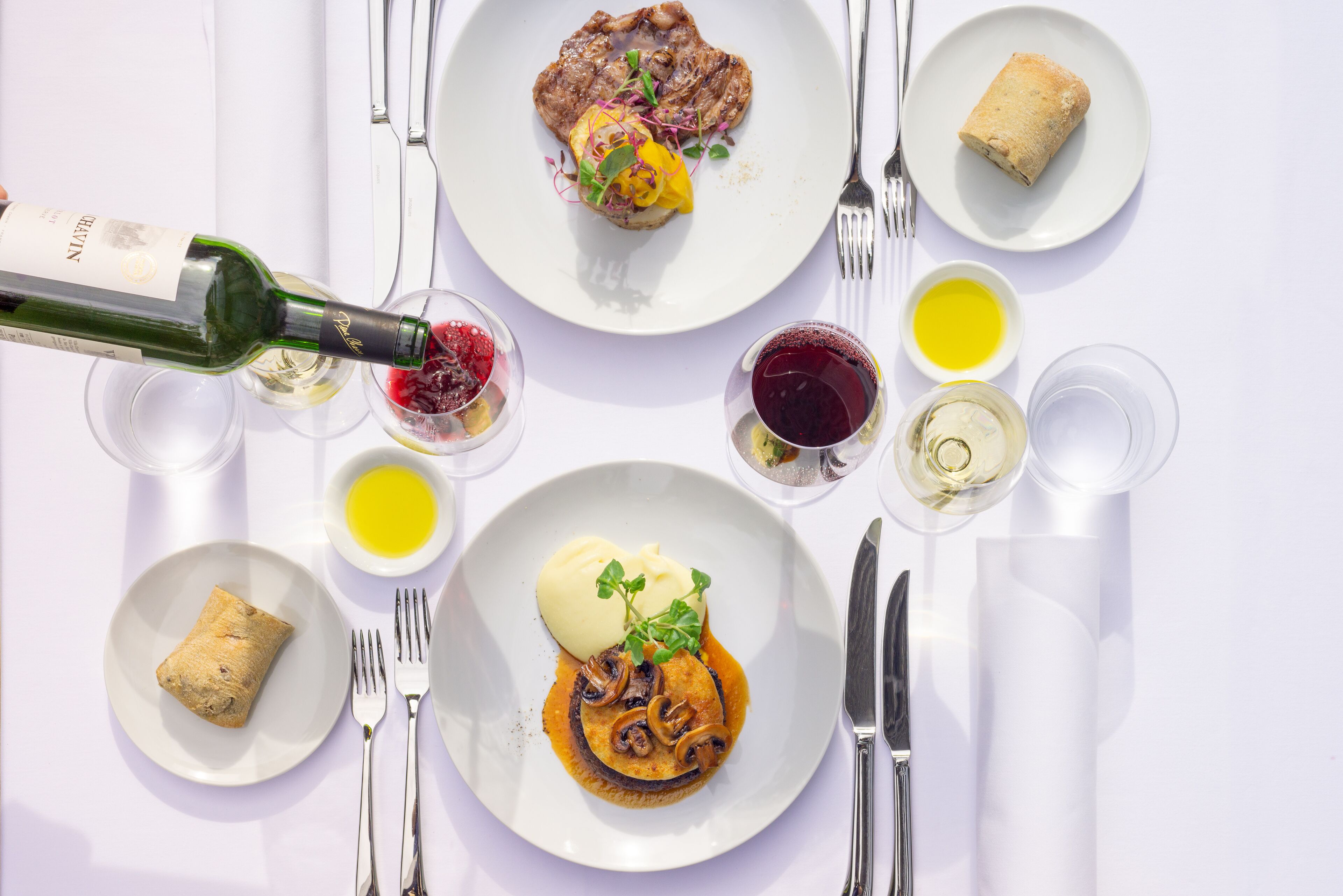 Ein Tisch ist reichhaltig gedeckt, mit verschiedenen Speisen, darunter ein Steak, ein Brötchen und eine Nachspeise, sowie mehreren Gläsern Wein und Saft.