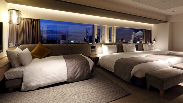 Una habitación de hotel moderna con vista a la ciudad, dos camas, un ventanal grande y una mesita de noche.