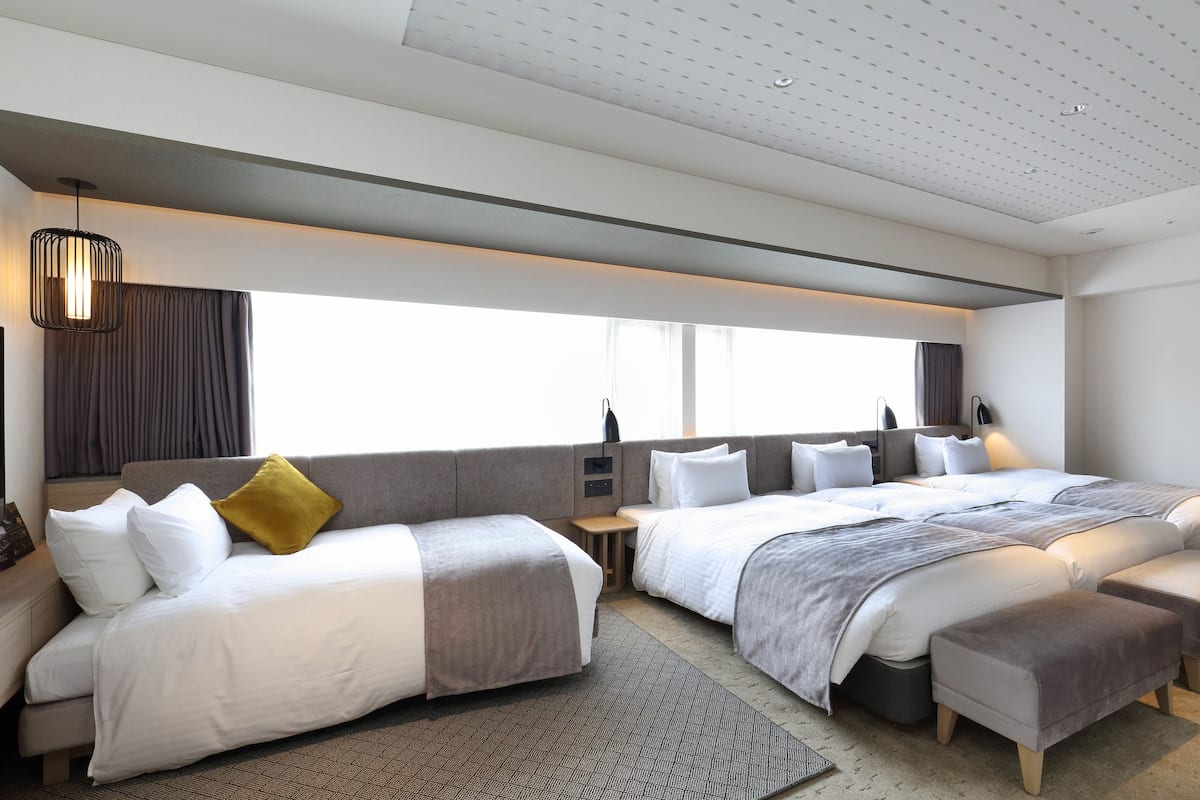 A modern hotel room with a large bed, a smaller sofa, a bedside table, and a window with curtains.