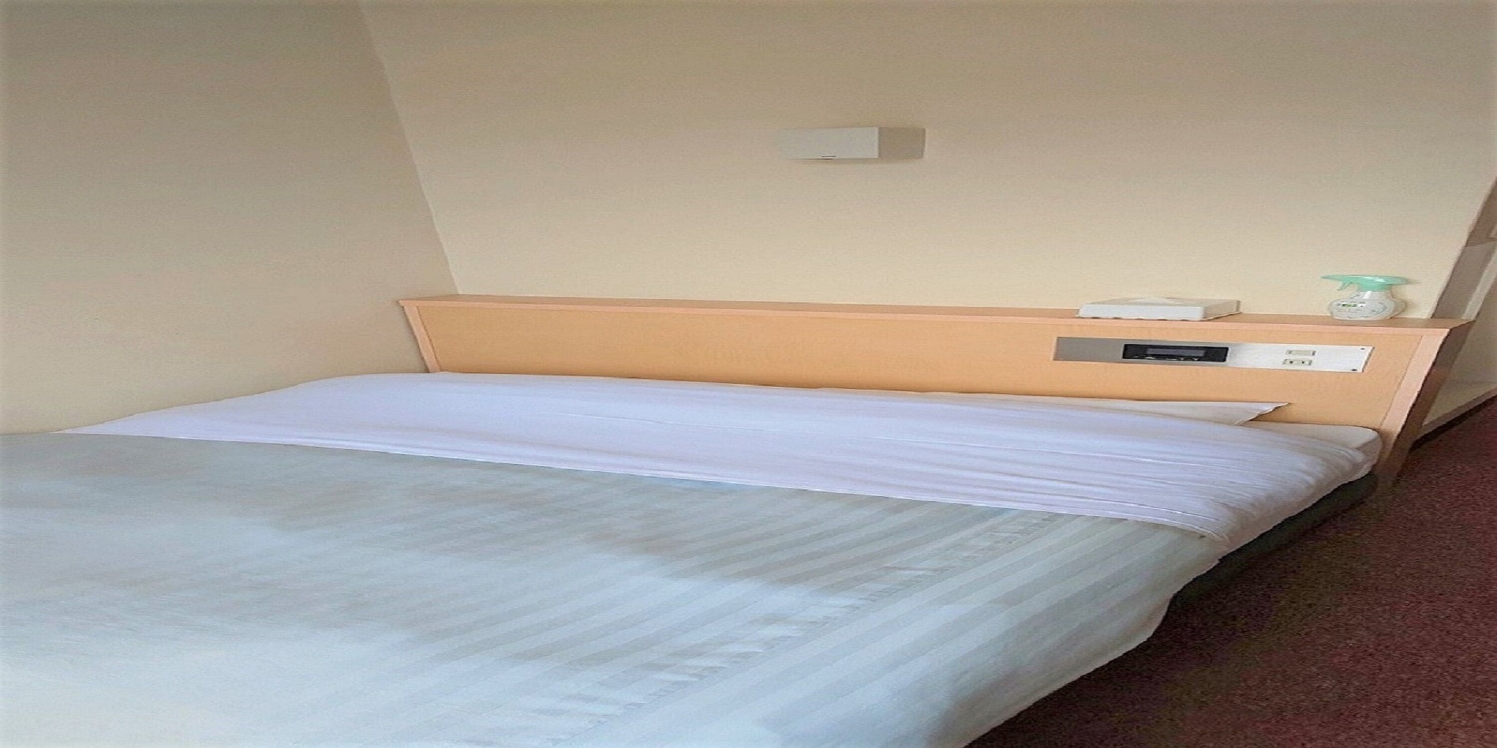 Een enkel bed met een houten hoofdbord, een witte matras en een klein nachtkastje met een lamp en een stopcontact.