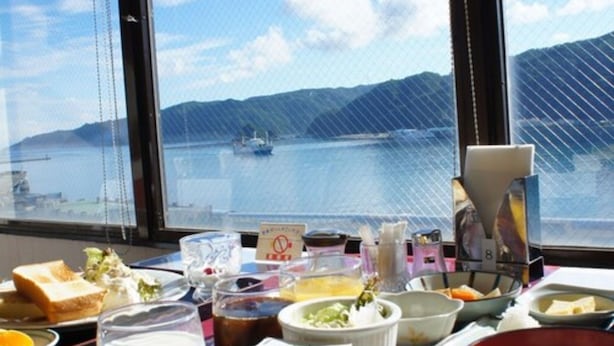 A table with various dishes and drinks, a window with a view of a body of water and mountains, and a sign indicating a no-smoking area. - Amami Port Tower Hotel (Amami)