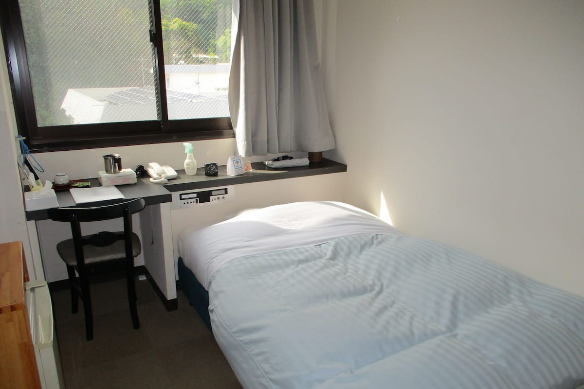 Una habitación pequeña y ordenada con una cama individual, un escritorio con silla, una ventana con vistas al exterior y un estante con varios objetos.