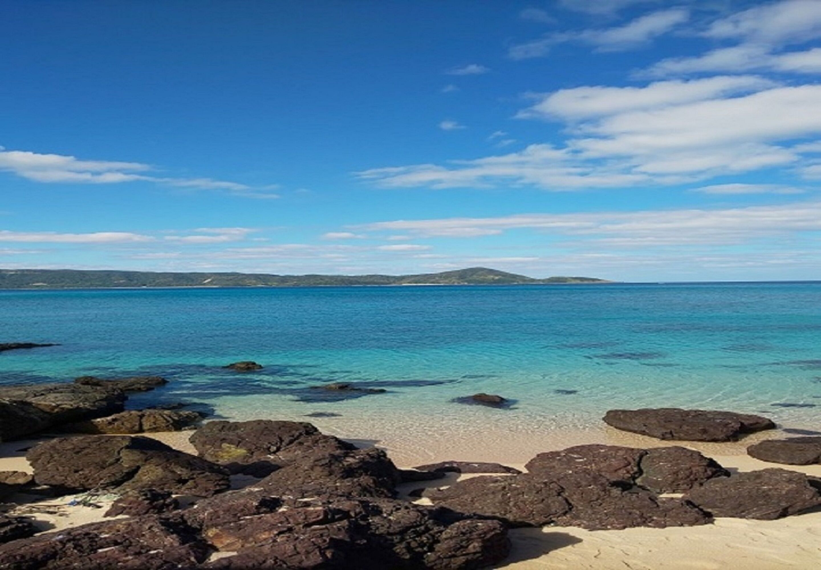 A beach with clear turquoise water, rocky foreground, and distant hills.