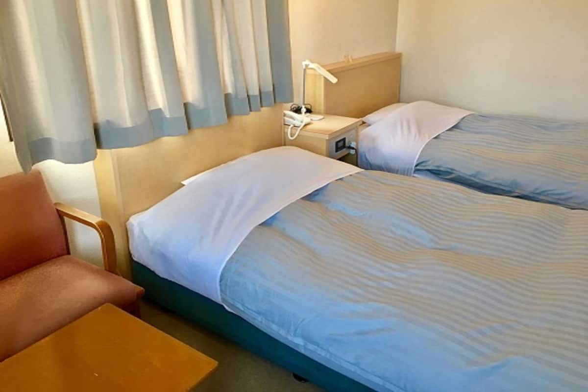 Habitación de hotel con dos camas, una silla, una mesita y una mesita de noche con lámpara.