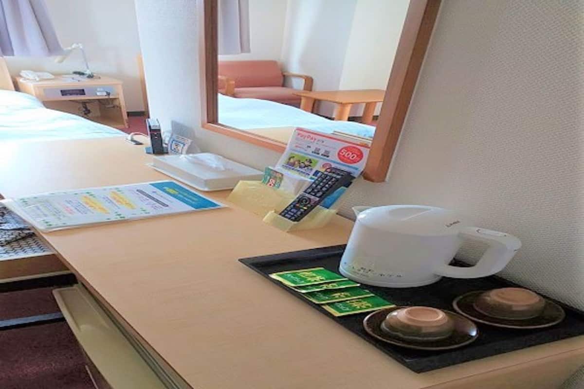 Habitación de hotel con escritorio, espejo, cama, mesita de noche, teléfono, cafetera y bandeja con tazas y tetera.