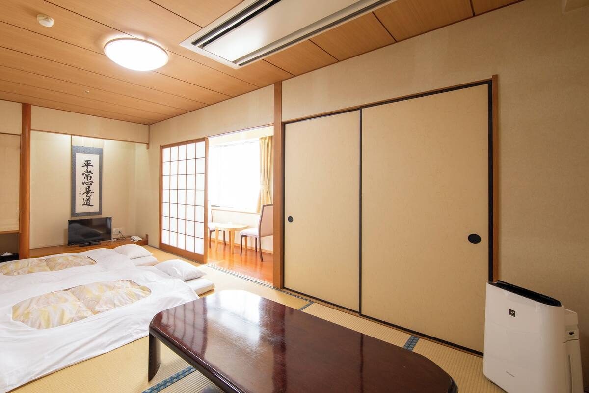 A traditional Japanese room with tatami flooring, a bed with white bedding, a television, a sliding door, and a ceiling fan.
