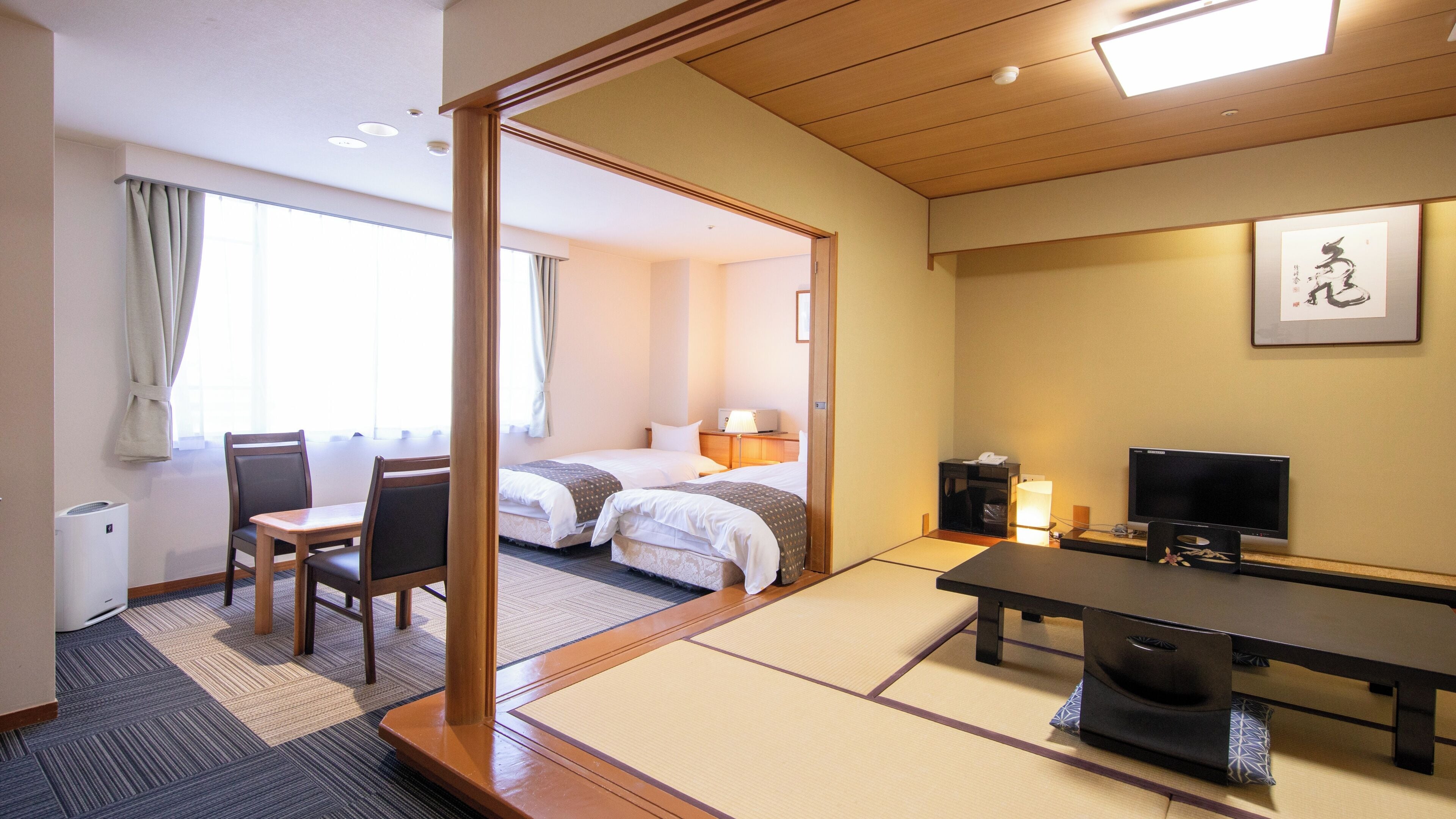 A hotel room with a traditional Japanese tatami mat floor, a bed, a desk with a chair, a television, and a bedside table.