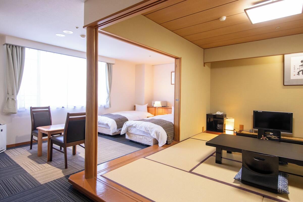 A hotel room with a traditional Japanese tatami mat floor, a bed, a desk with a chair, a television, and a bedside table.
