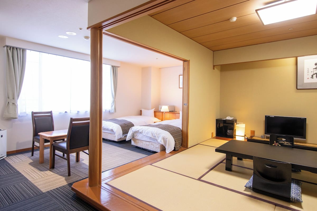 Ein Hotelzimmer mit traditionellem japanischem Tatami-Boden, einem Bett, einem Schreibtisch mit Stuhl, einem Fernseher und einem Nachttisch.